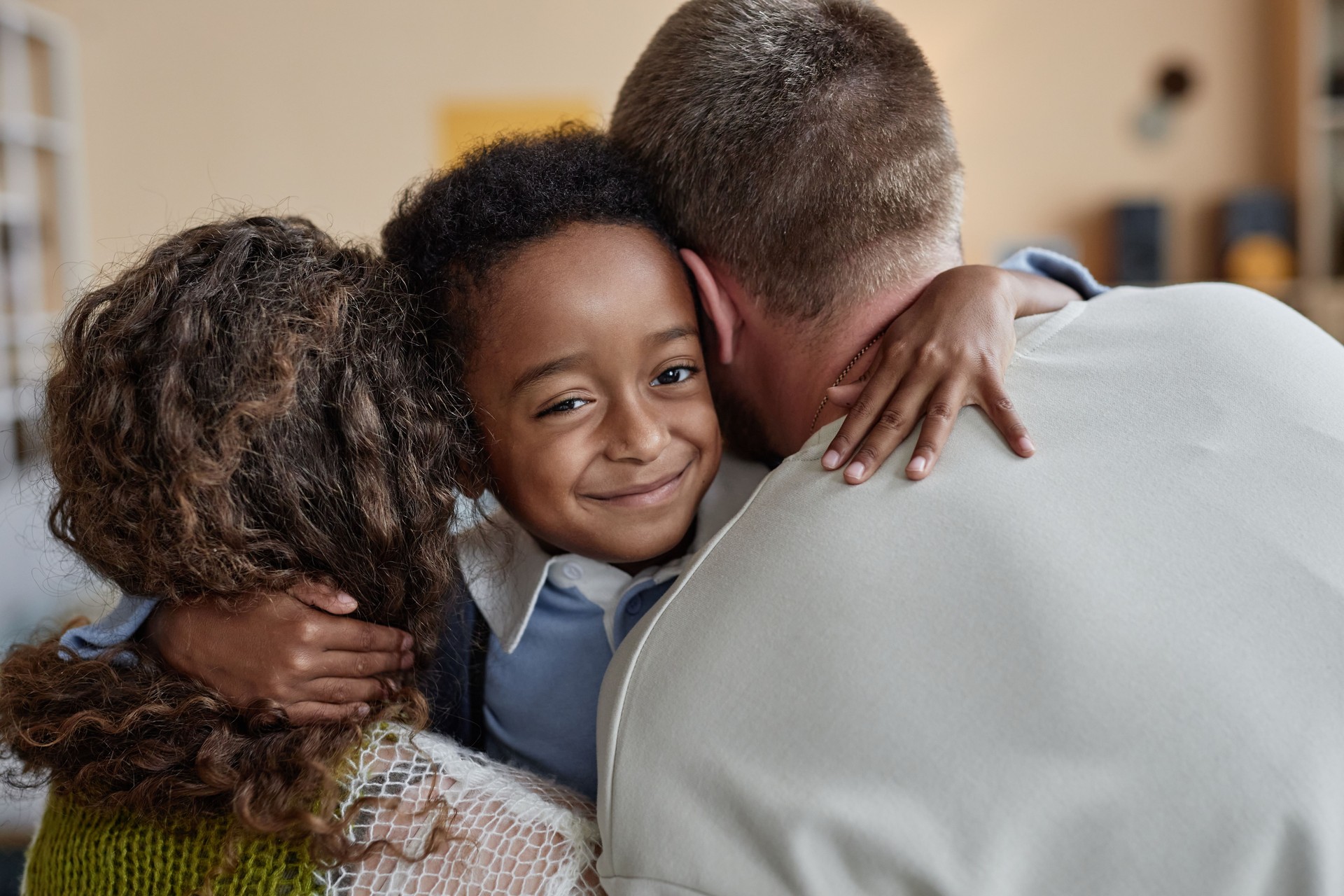 Smiling Black Boy Looking at Camera while Hugging Adoptive Parents Indoors